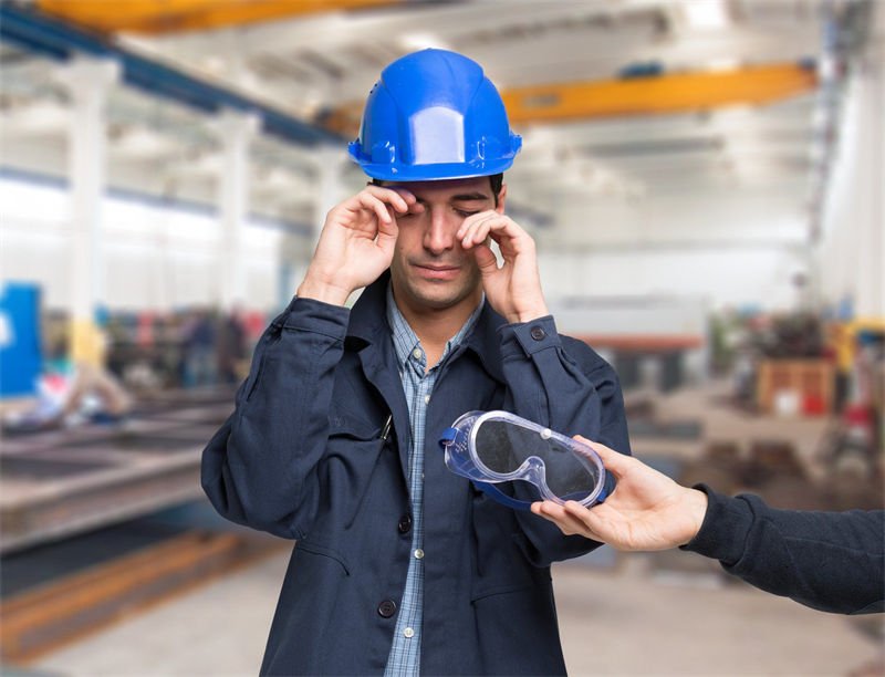 A worker wearing a blue hard hat rubbing his eyes while another person hands him safety goggles in an industrial setting