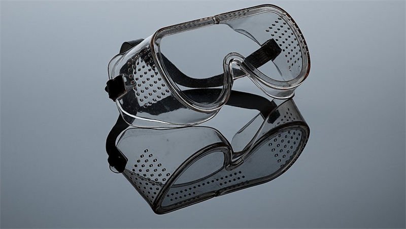 Transparent laser safety goggles with perforated ventilation and an adjustable strap, placed on a reflective surface.