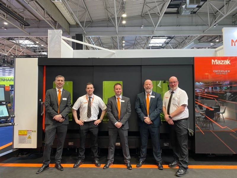 Mazak team posing in front of the OPTIPLEX 3015 NEO laser cutter at an exhibition