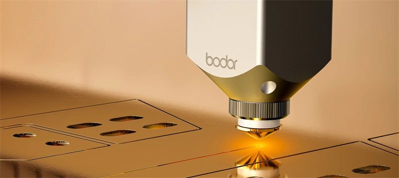 A Bodor laser cutting head performing precision cuts on a golden metallic surface.