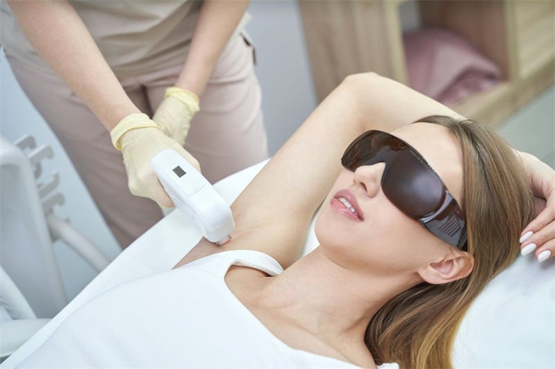 A woman wearing protective laser safety glasses undergoing a laser hair removal procedure by a professional in a clinic