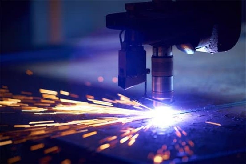 Plasma cutting machine head emitting a bright plasma arc and sparks while cutting through metal in a dimly lit workshop=