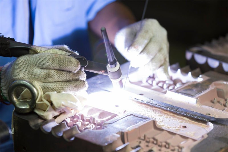 Worker performing TIG welding on a detailed metal component with precision tools and protective gloves