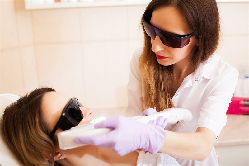 A beauty professional wearing protective laser goggles performing a cosmetic laser treatment on a client, who is also wearing safety glasses