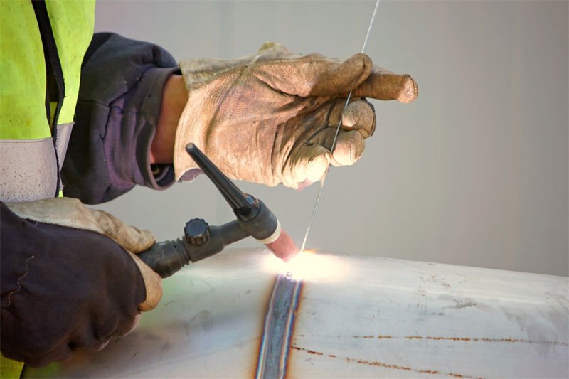 A welder performing TIG welding on a cylindrical pipe with protective gloves and a filler rod