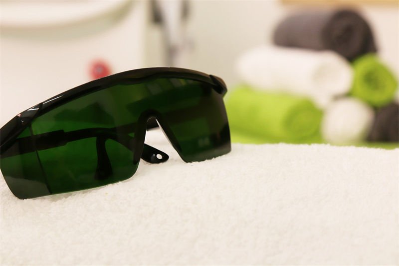 UV Laser Safety Glasses for Eye Protection A pair of green-tinted UV laser safety glasses placed on a white towel in a spa-like setting with folded towels in the background