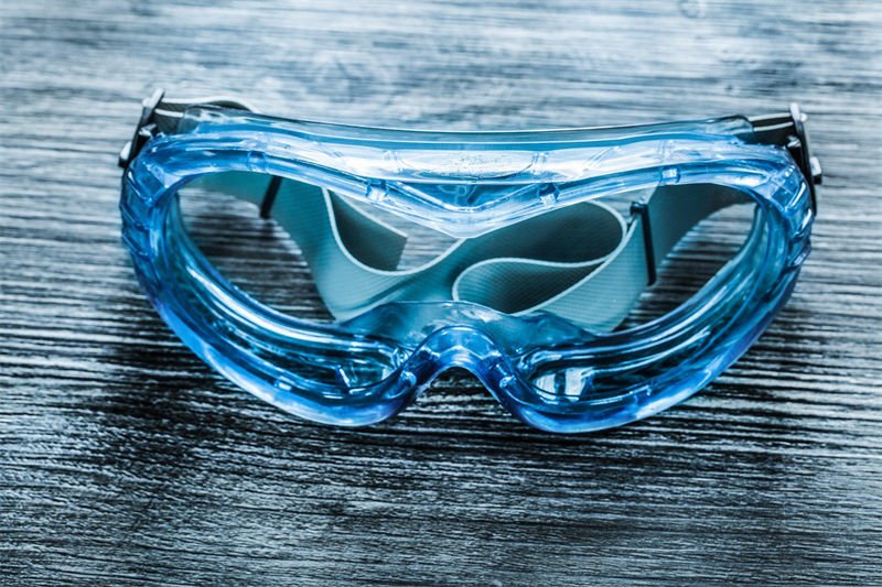 A pair of blue-tinted protective goggles with an adjustable strap placed on a textured wooden surface