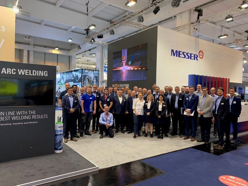 A large group photo of the Messer team at an exhibition booth showcasing welding and cutting solutions