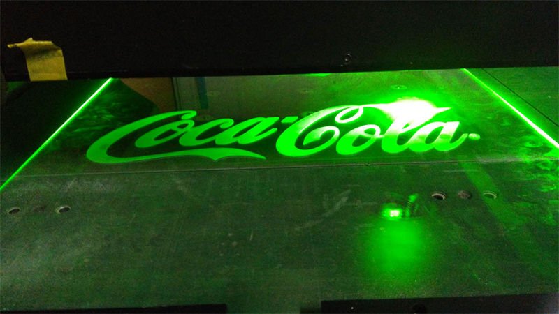 Laser engraving a Coca-Cola logo on a glass panel with green laser light.