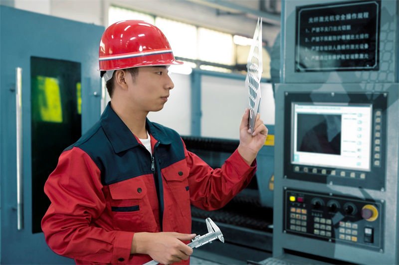 A worker in a red uniform and safety helmet inspects a freshly laser-cut stainless steel sheet