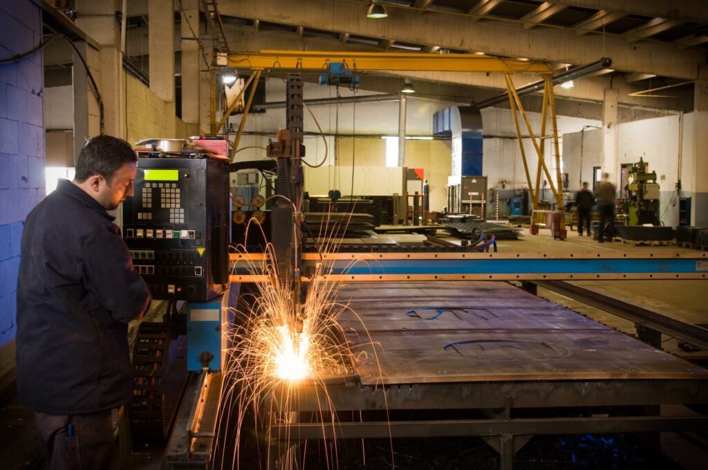 A worker monitors a plasma cutting machine as it cuts through a large metal sheet, producing bright sparks in an industrial setting.