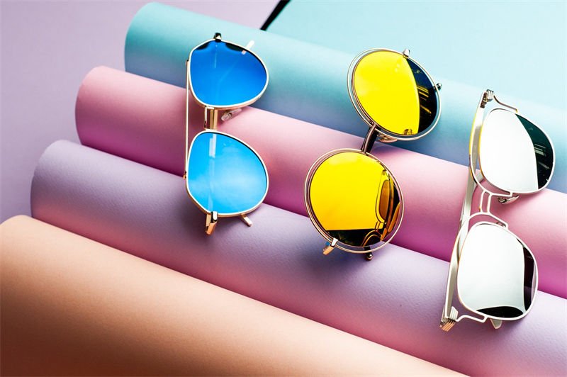 A collection of UV protection sunglasses with colorful lenses in blue, yellow, and reflective silver displayed on pastel-colored rolls