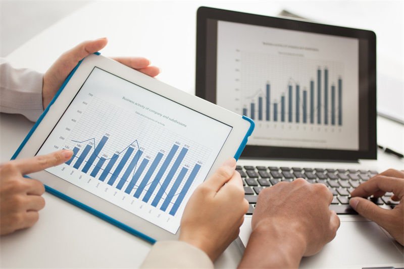 Business professionals analyzing bar chart data on a tablet and laptop screen, discussing trends and market dynamics