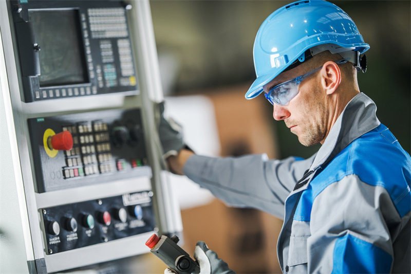 A technician wearing a blue helmet and protective glasses inspecting a control panel in an industrial setting