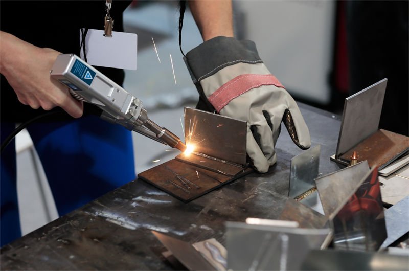 A skilled worker using a handheld laser welding tool on small metal parts at a workstation.