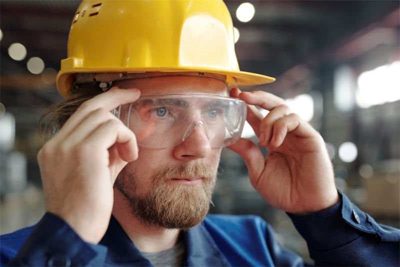 A male worker adjusting safety goggles while wearing a yellow hard hat in an industrial setting