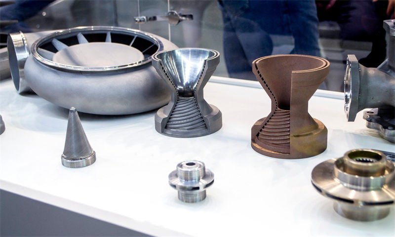 A collection of 3D-printed metal parts, including turbine components and conical structures, displayed on a white table